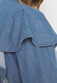 Jeansjacke mit Rüschkragen, hellblauem Material und strukturiertem Design. Besitzt sichtbare Nähte und einen weichen, texturierten Stoff.