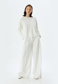 Vit stickad tröja med rund halsringning i kombination med lösa vita byxor. Outfiten har rena linjer och en elegant, minimalistisk design.