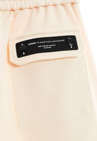 Una prenda de color melocotón con una etiqueta negra. La etiqueta incluye texto que indica la colaboración de la marca y el origen, con dos botones negros.