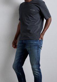 Mörkgrå t-shirt med kort ärm och rund hals, med en liten vit logotyp. Bärs tillsammans med blå slim-fit jeans som har ljusa blekningar.