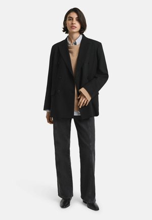Black double-breasted coat over a tan sweater, light blue shirt, and dark jeans. Outfit completed with black pointed ankle boots.