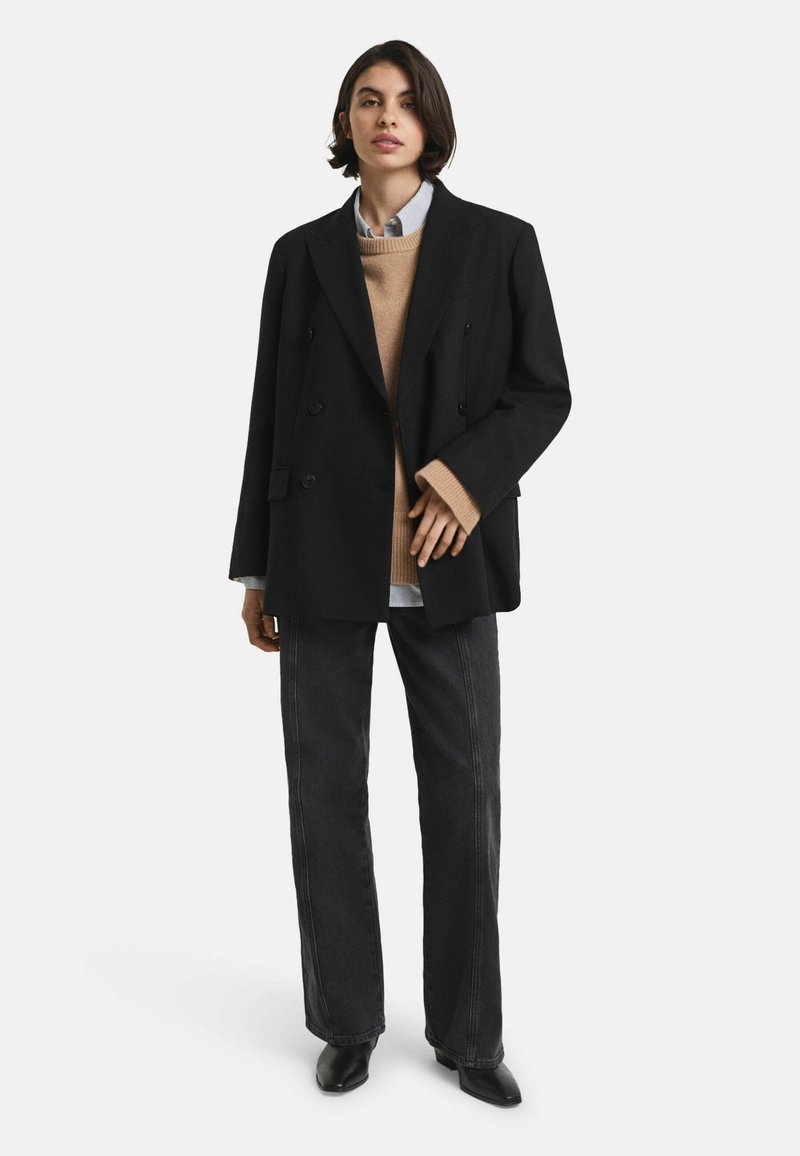 Manteau noir à double boutonnage porté sur un pull beige, une chemise bleu clair et un jean foncé. Tenue complétée par des bottines noires à talons aiguilles.