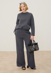Kvinde med kort blondt hår iført mørkegrå sweatshirt og bukser med vide ben, holder en lille tekstureret håndtaske og har hælede sandaler på.