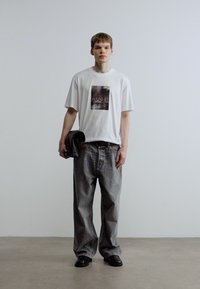 Άσπρο κοντομάνικο μπλουζάκι με γραφικό τύπωμα, σε συνδυασμό με άνετο γκρι τζιν παντελόνι και μαύρα παπούτσια, στέκεται μπροστά σε ουδέτερο φόντο.