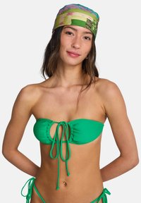 Πράσινο τοπ μαγιό με άνοιγμα σε σχήμα κλειδαρότρυπας και ρυθμιζόμενα κορδόνια. Λείο υλικό, χωρίς τιράντες. Το μοντέλο φοράει  πολύχρωμο μαντήλι με σχέδια.