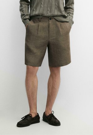 Uomo che indossa pantaloncini marroni su misura, maglione grigio lavorato a trecce e scarpe nere con lacci, in piedi con le mani in tasca.