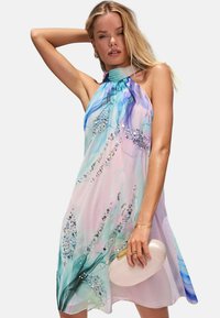 Robe à encolure halter en tissu transparent, avec un dégradé pastel, des motifs floraux et des ornements. Assortie à un sac à main de couleur claire.
