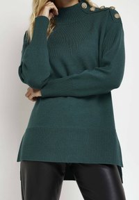 Grön stickad tröja med hög ribbad krage, sidofragor och dekorativa guldknappar på axeln; matchad med svarta läderbyxor.