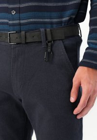 Black fabric belt with a plastic buckle, attached carabiner clip, paired with dark navy textured pants and a multi-striped shirt.