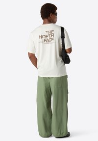 Homme portant des lunettes de soleil, t-shirt blanc "The North Face", pantalon vert ample et sac en bandoulière noir, se tenant de dos contre un fond blanc.