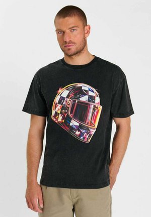 Uomo che indossa una maglietta nera con grafica colorata a scacchi di casco da corsa, in piedi su sfondo bianco uniforme con espressione neutra.