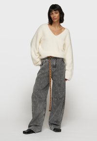 Suéter de gran tamaño, color crema, con escote en v y puños acanalados, combinado con unos jeans de pierna ancha de patrón gris claro y un cinturón marrón. Zapatos negros completan el look.