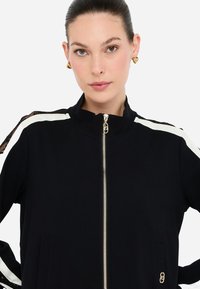 Kvinde med glat tilbageholdt hår, iført guld hjerteformede øreringe og en sort zip-up jakke med hvide striber og blonder på ærmerne.