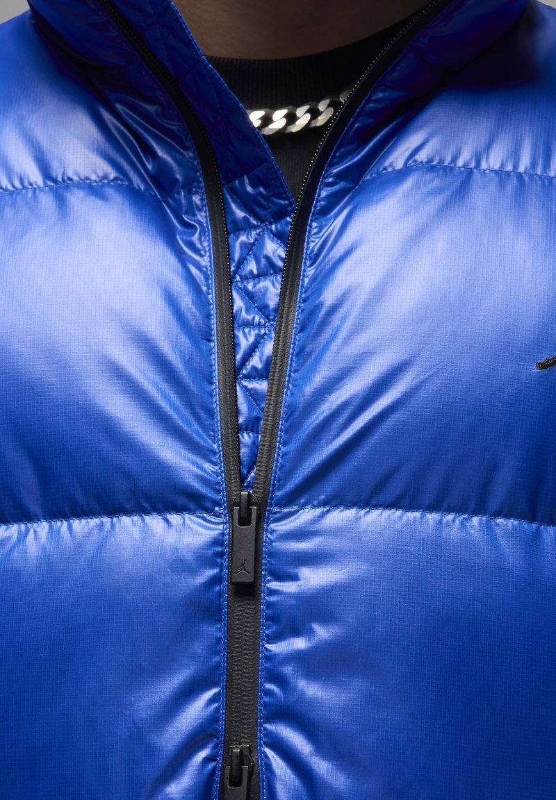 Chaqueta de plumas azul con un acabado brillante y acolchado, cierre de cremallera completo y herrajes negros. Presenta un logo en la parte superior izquierda.