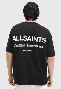 Melnas kokvilnas t-krekls ar izteiksmīgu baltu tekstu "ALLSAINTS" un sarežģītu rakstu uz muguras. Īsas piedurknes un brīvs griezums.