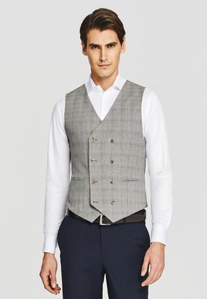 Mężczyzna w szarej kraciastej kamizelce dwurzędowej na białej koszuli i granatowych spodniach, stojący na jednolitym tle.