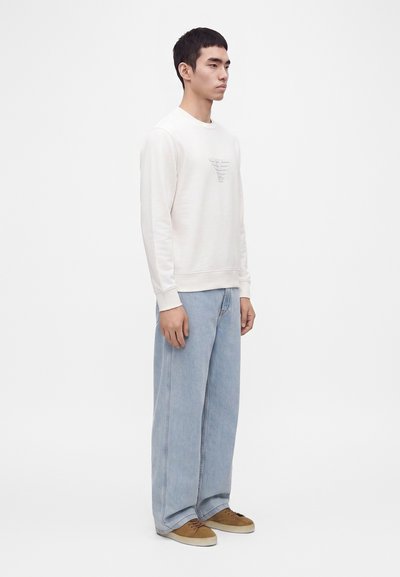Un jeune homme se tient debout, portant une chemise blanche à manches longues avec un logo discret, un jean large bleu clair et des baskets marron sur un fond blanc.