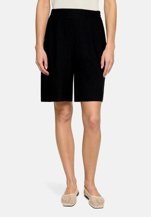 Kobieta w czarnych, dzianinowych szortach do kolan i beżowych plecionych butach typu slip-on na jednolitym tle.