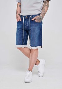 Jeans kratke hlače v temno modri barvi z belim vrvico in zvitimi manšetami. Kombinirane z belimi supergami. Imajo vidne šive in žepe.