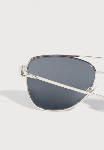 Γυαλιά ηλίου με ασημένιο σκελετό και σκουρόχρωμους γκρι ορθογώνιους φακούς που ακουμπάνε σε λευκή επιφάνεια, με έναν φακό και το λεπτό μπράτσο εστιασμένα.