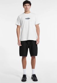 Valge puuvillane T-särk sinise logoga, mustade teksapükstega, esitades lõdva lõikega. Modell kannab musti jalanõusid ja valgeid sokke.