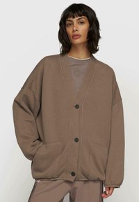 Brauner oversized Cardigan mit V-Ausschnitt, einem Knopfverschluss und seitlichen Taschen. Hergestellt aus weichem Stoff und mit lockerer Passform. Über einem grauen Oberteil getragen.