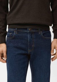 Mann trägt dunkelblaue Jeans und einen schwarzen Pullover, die Hände entspannt neben dem Körper, Fokus auf Taille und oberem Oberschenkelbereich.