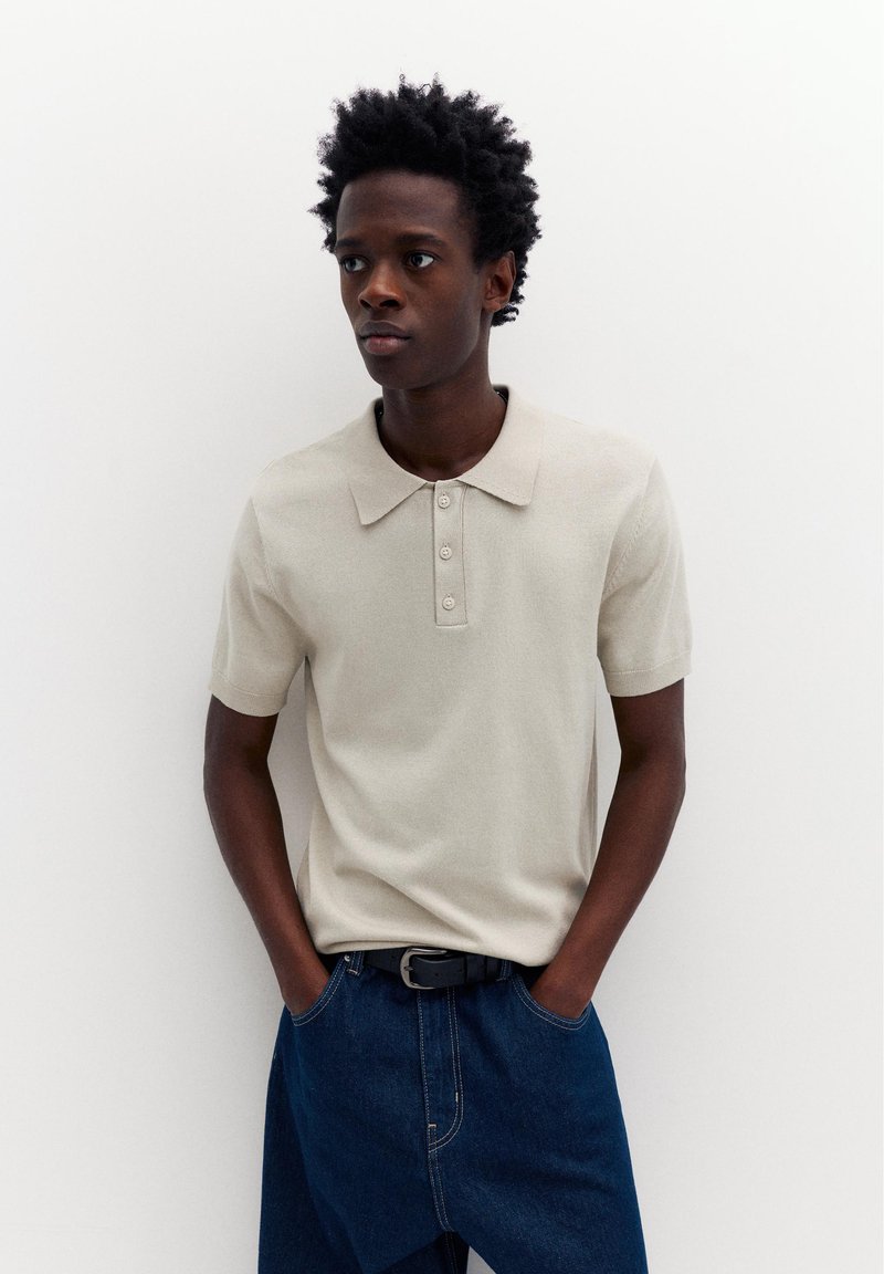 Giovane uomo con capelli naturali che indossa una polo beige a maniche corte infilata in un paio di jeans blu scuro, in piedi con le mani nelle tasche.