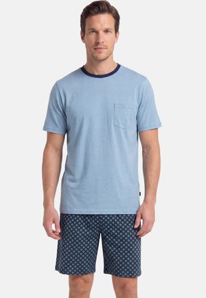 Mężczyzna w jasnoniebieskim T-shircie z krótkim rękawem i kieszenią z przodu oraz w ciemnoniebieskich, wzorzystych szortach, stojący na białym tle.