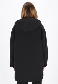 Donna che indossa un cappotto nero con cappuccio, vista di spalle, in piedi contro uno sfondo bianco uniforme.