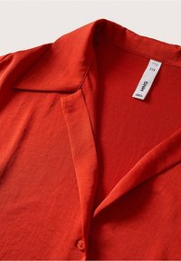 Chemise à col rouge vif avec un bouton visible, comportant une étiquette blanche indiquant la taille et les instructions d'entretien à l'intérieur de l'encolure.