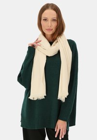 Beżowy szalik luźno zarzucony na szyi, wykonany z miękkiego materiału. Stylizowany z ciemnozielonym, dużym swetrem z dzianiny, z prążkowanymi wykończeniami.