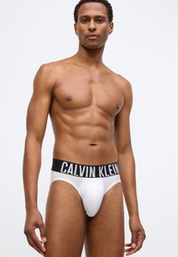 Λευκά σλιπ με μαύρη ζώνη "CALVIN KLEIN", που προβάλλουν μια λεία υφή υφάσματος και εφαρμοστή σχεδίαση. Το μοντέλο ποζάρει από μπροστά.