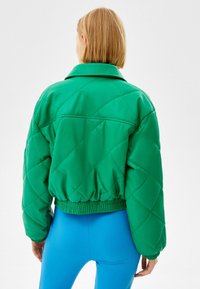Zelená prošívaná bunda s límcem a elastickým lemem, zobrazená ze zadní strany. Lehká tkanina, spojená se světlomodrými legínami.