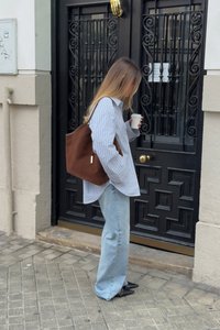 Brūns zamšs tote soma, svītrainā gaiši zila un balta krekls, gaiši zili džinsi, turot kafijas tasi, stāvot priekšā melnām durvīm ar dekoratīviem režģiem.