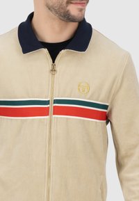 Beige Zip-Jacke mit Reißverschluss und marineblauem Kragen, versehen mit horizontalen Streifen in Rot, Grün und Weiß. Goldenes Logo auf der Brust gestickt.