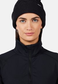 Berretto a maglia nero con un motivo testurizzato, indossato su una giacca nera a collo alto con cerniera, tessuto liscio e design aderente.