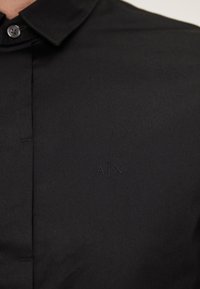 Camisa negra de botones con una textura suave, que presenta un cuello simple y un logo en relieve en el pecho.