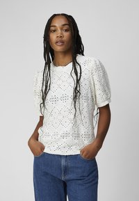 Witte top met pofmouwen, intricaat bloemen-ogen patroon, zachte textuur, ronde halslijn en een relaxte pasvorm, gecombineerd met blauwe denim jeans.