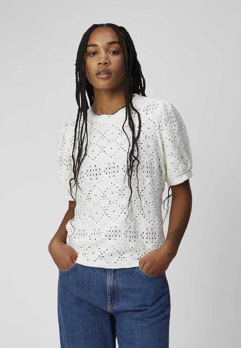 Witte top met pofmouwen, intricaat bloemen-ogen patroon, zachte textuur, ronde halslijn en een relaxte pasvorm, gecombineerd met blauwe denim jeans.