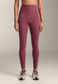 Donna che indossa leggings color malva a vita alta e scarpe da ginnastica bianche, in piedi contro uno sfondo neutro e semplice.