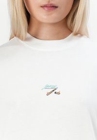 Balta medvilninė marškinėlių su apvaliu kaklu, turinti spalvingą siuvinėtą logotipo dizainą ant krūtinės. Lygi tekstūra ir laisvas fasonas.