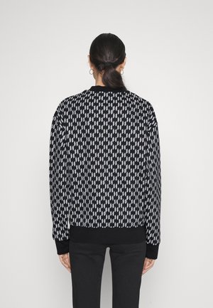 Sweat-shirt noir et blanc à motifs présentant un design géométrique répétitif, avec des poignets et un col côtelés. Porté avec un pantalon noir.