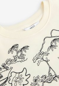 Tissu couleur crème avec motifs floraux et feuillages brodés en noir près d'un col rond et une étiquette "NEXT" partiellement visible.