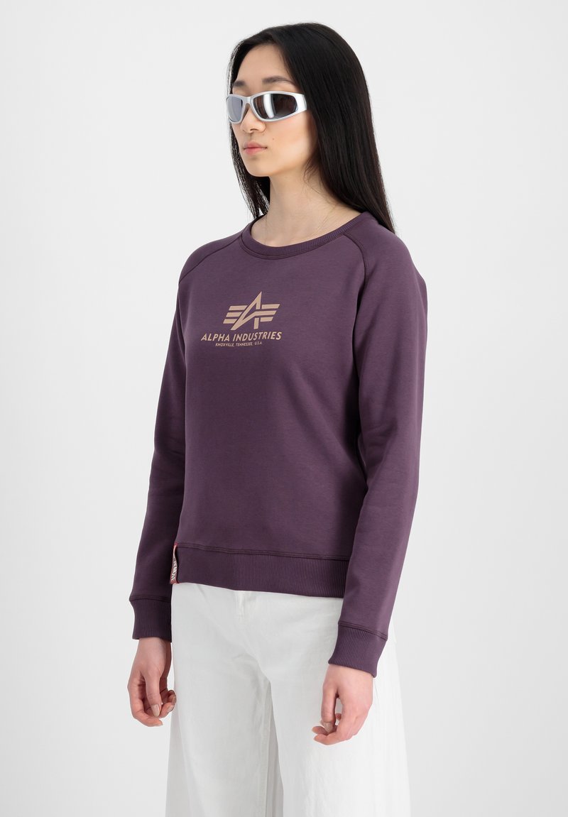 Lilla sweatshirt fra Alpha Industries med rund halsudskæring, rib strik ved håndleddene og et guld logo i midten; båret med hvide bukser.