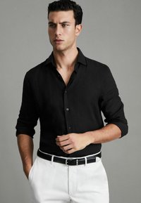 Schwarzes Leinenhemd mit langen Ärmeln, einem geknöpften Kragen und hochgekrempelten Ärmeln, kombiniert mit weißen Hosen und einem schwarzen Gürtel mit einer silbernen Schnalle.