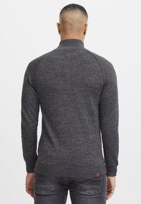 Mann mit kurzen Haaren, der einen dunkelgrauen, gerippten Pullover und dunkelgraue Jeans trägt, von hinten vor einem schlichten weißen Hintergrund gezeigt.