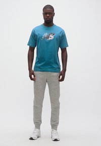 Άνδρας που στέκεται φορώντας γαλάζιο μπλουζάκι New Balance, γκρι φόρμες και λευκά αθλητικά παπούτσια μπροστά σε λευκό φόντο.