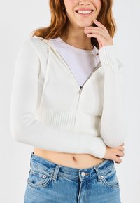 White cropped cardigan with a front zipper, ribbed cuffs, and a knitted texture, paired with high-waisted denim jeans.