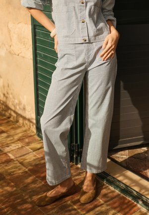 Personne portant une tenue rayée bleu et blanc à boutons avec poches, debout en plein air sur des dalles en pierre, chaussée de chaussures plates marron.